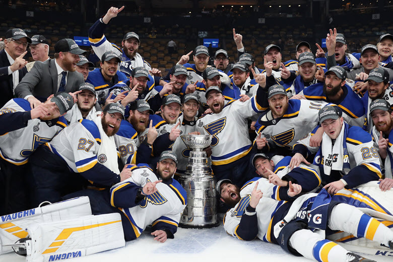 The St. Louis Blues pose with the 2019 Stanley Cup while sports fans cheer.