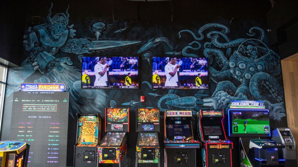 Classic arcade games line the wall, which features a mural, at 4 Hands at The District.