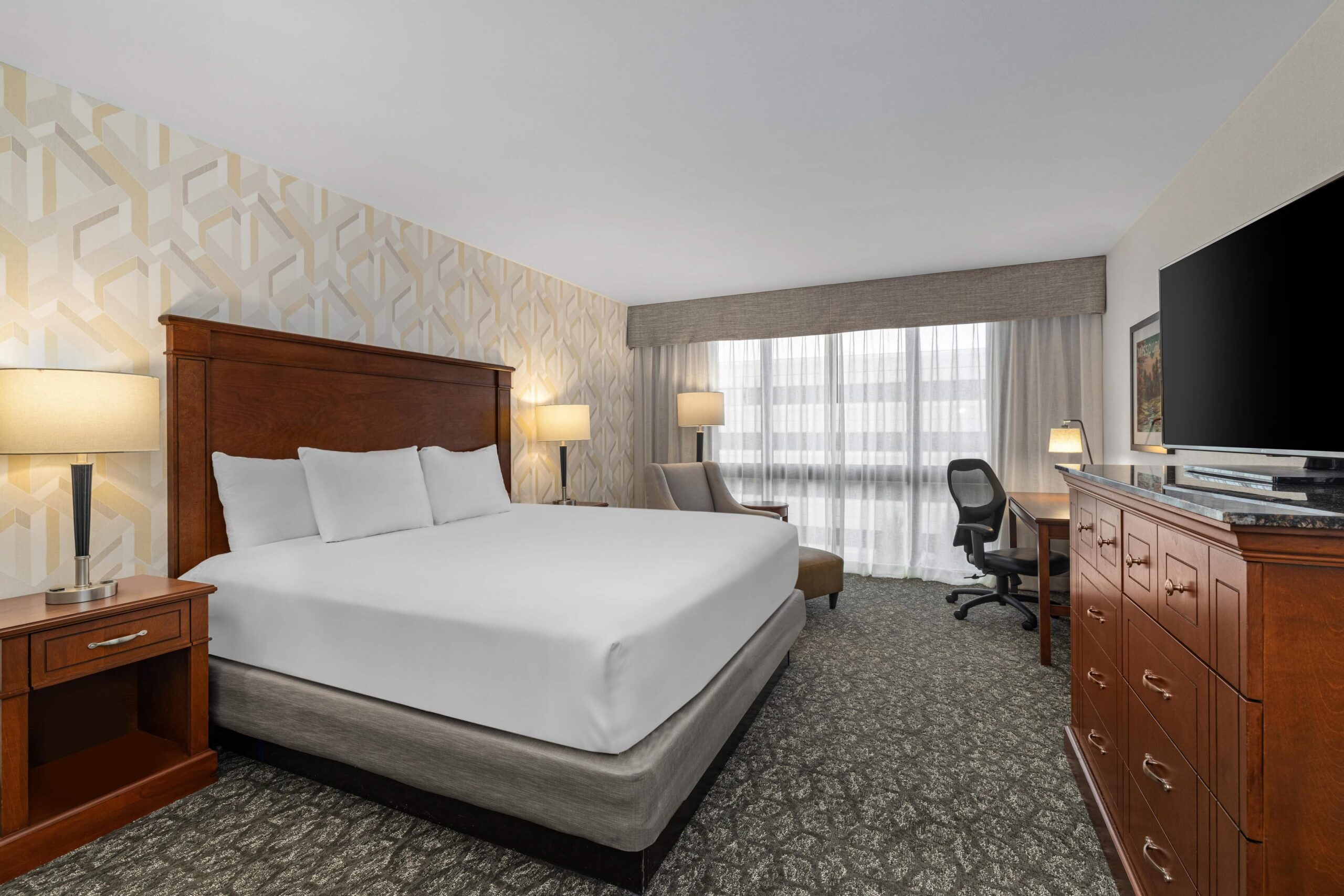 Spacious guest room featuring a comfortable king-sized bed, an ergonomic work desk with a chair, and a flat-screen TV on a wooden dresser at Drury Inn & Suites St. Louis Creve Coeur.