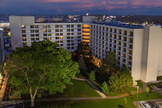 Exterior dusk view of the Hilton St. Louis Airport hotel building featuring a lush green courtyard with large trees and the airport terminal in the background.
