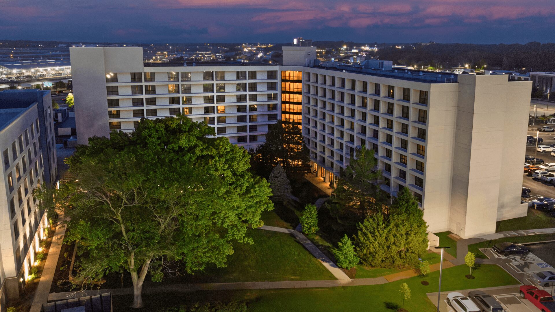 Exterior dusk view of the Hilton St. Louis Airport hotel building featuring a lush green courtyard with large trees and the airport terminal in the background.