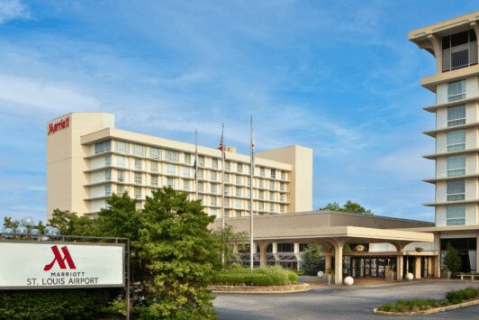 Exterior view of the Marriott St. Louis Airport hotel illuminated at dusk, featuring a modern architectural facade and the brightly lit Marriott logo against a clear evening sky.