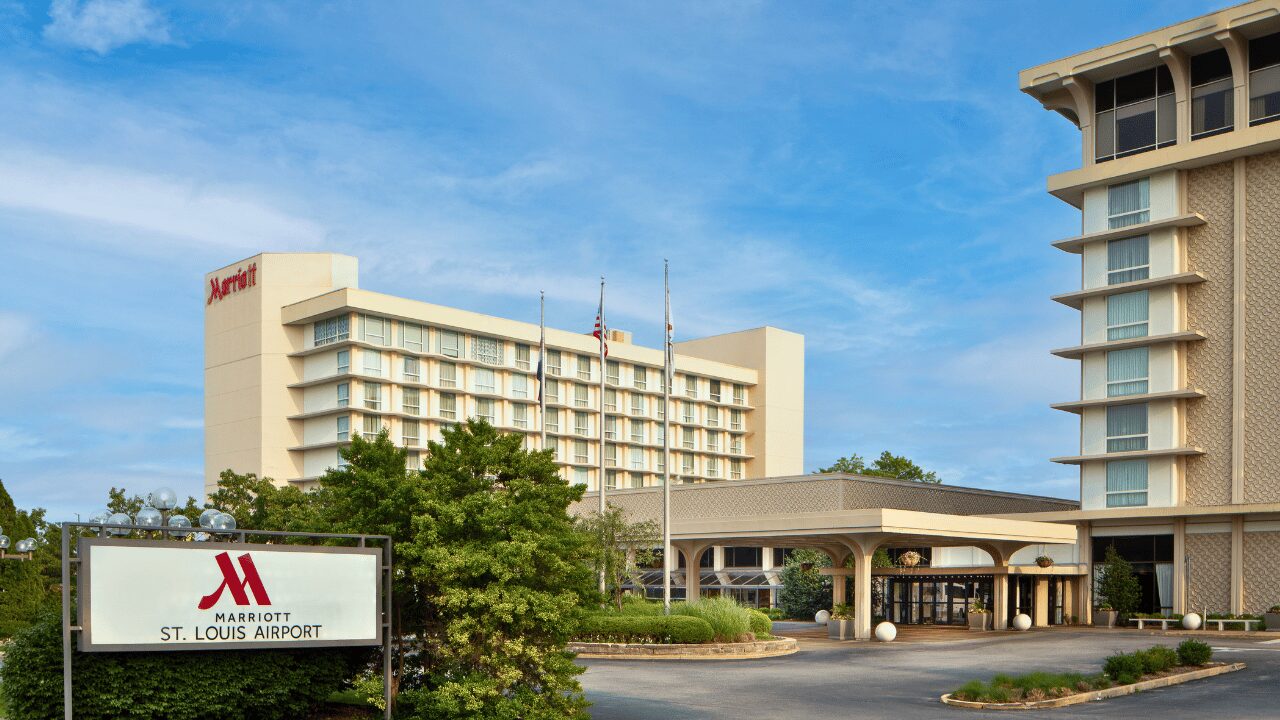 Exterior view of the Marriott St. Louis Airport hotel illuminated at dusk, featuring a modern architectural facade and the brightly lit Marriott logo against a clear evening sky.