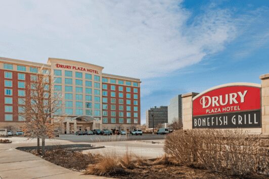 Aerial view of the modern, multi-story exterior and adjacent covered parking garage of the Drury Plaza Hotel St. Louis Brentwood on a bright, sunny day.