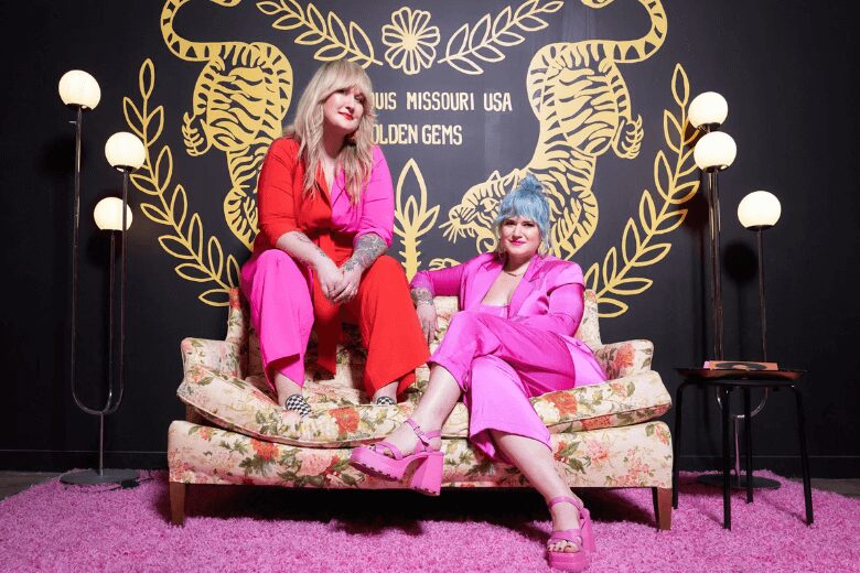 Founders Susan Logsdon and Amanda Helman of Golden Gems sitting on a floral vintage sofa in front of a black wall with a gold tiger mural in Midtown Alley, St. Louis.