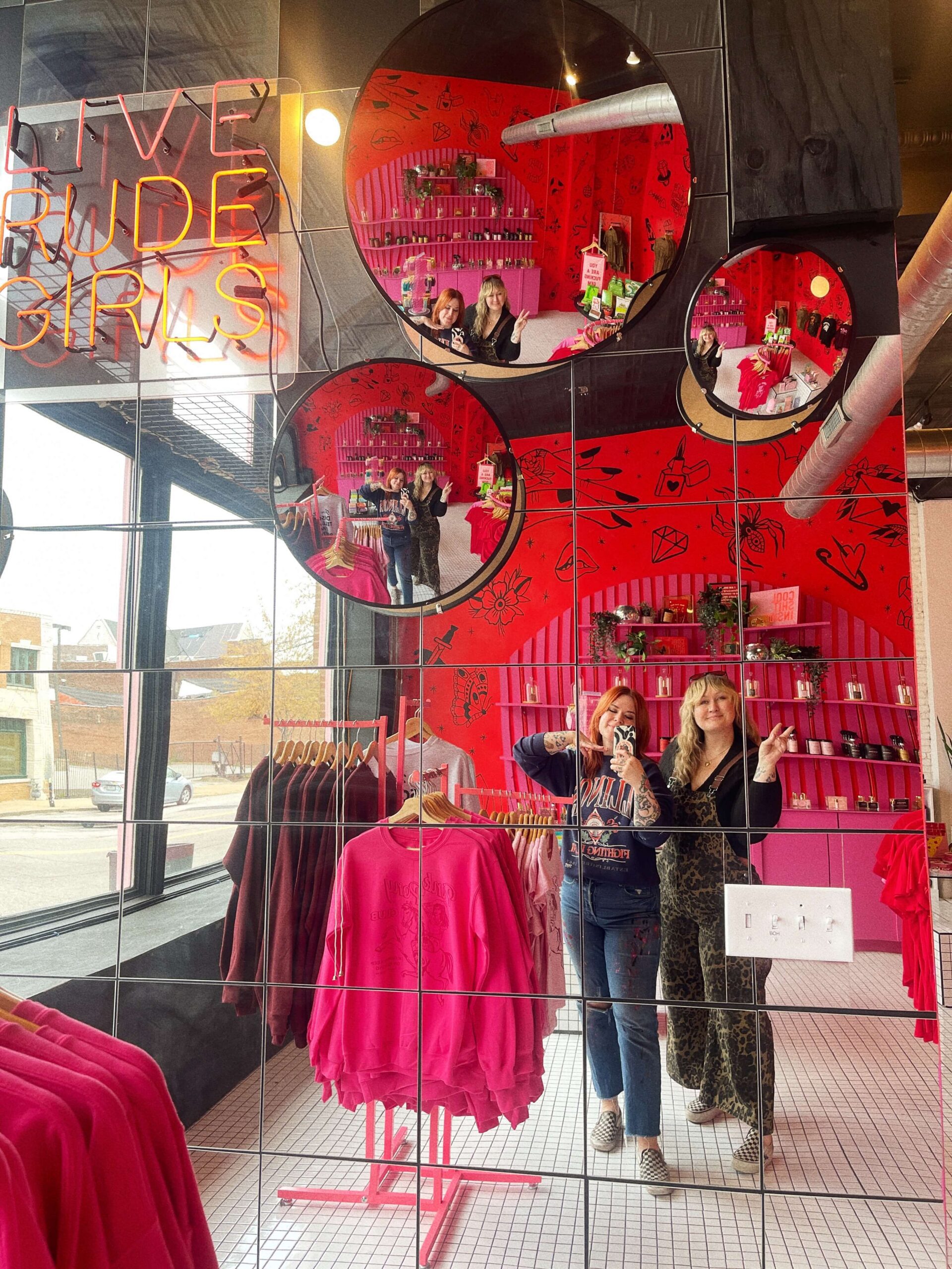 A multi-panel grid mirror in the Golden Gems shop reflecting pink and maroon sweatshirts and a "Live Rude Girls" neon sign, perfect for customer social media photos.