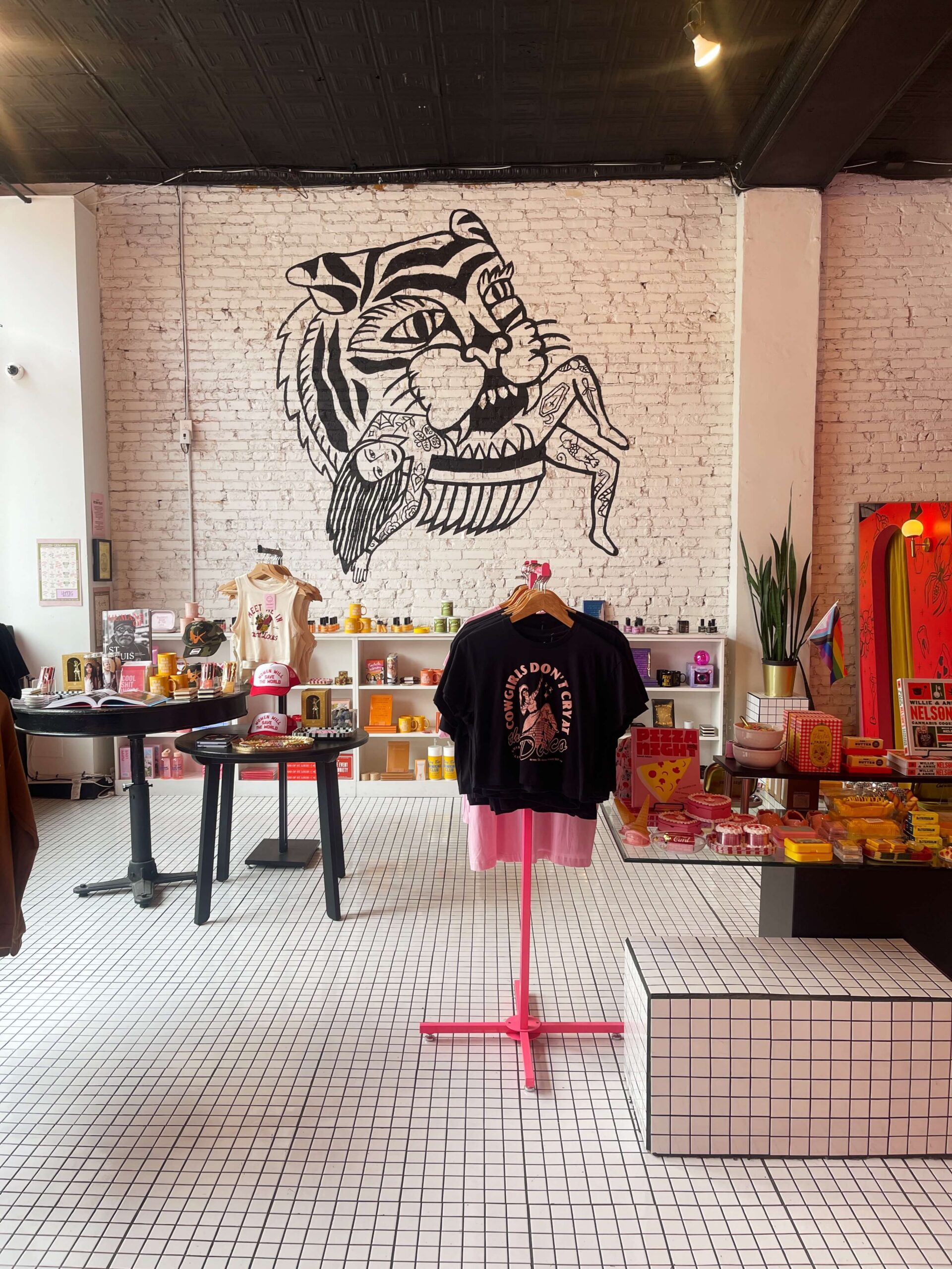 A large black and white tiger mural on a white brick wall inside Golden Gems Midtown Alley, overlooking racks of feminist graphic t-shirts and lifestyle accessories.